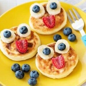 Três panquecas douradas decoradas de forma lúdica com fatias de banana, mirtilos e framboesas, formando rostos divertidos.
