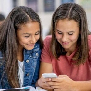 Duas meninas sentadas uma ao lado da outra na sala de aula, sorrindo e olhando para um celular Duas meninas sentadas uma ao lado da outra na sala de aula, sorrindo e olhando para um celular