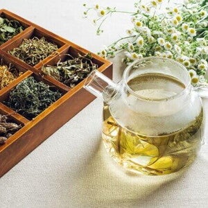 Uma chaleira de vidro com chá de ervas amarelado ao lado de uma caixa de madeira com compartimentos cheios de diversas ervas secas e flores brancas de camomila.