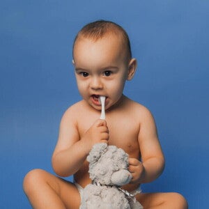 Bebê sentado com uma escova de dentes na boca, segurando um urso de pelúcia Bebê sentado com uma escova de dentes na boca, segurando um urso de pelúcia
