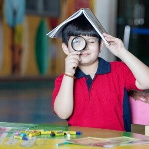 Um menino sorridente em uma mesa colorida, equilibrando um livro na cabeça e olhando através de uma lupa.