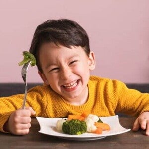 Menino pequeno sorrindo com entusiasmo ao segurar um garfo com brócolis. À sua frente, um prato com brócolis, cenoura e couve-flor ilustra como montar um prato saudável infantil de forma atrativa e nutritiva para crianças.