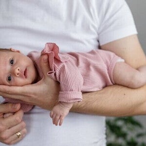 Close-up de um pai segurando um recém-nascido com a barriga para baixo em seu antebraço, em uma posição de "avião".