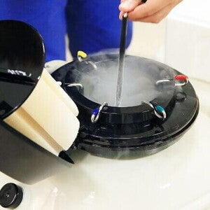 Close-up de um tanque de nitrogênio líquido aberto, com vapor frio subindo. Uma pessoa está inserindo ou retirando uma palheta ou recipiente de armazenamento de embriões.