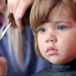 Uma menina recebendo um corte de cabelo. Ao seu lado, a mão de uma mulher segura uma mecha do seu cabelo, enquanto outra mão segura uma tesoura pequena Uma menina recebendo um corte de cabelo. Ao seu lado, a mão de uma mulher segura uma mecha do seu cabelo, enquanto outra mão segura uma tesoura pequena