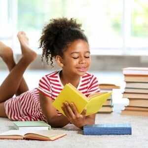 Uma menina sorridente, está deitada de bruços em um tapete, lendo um livro. Vários outros livros estão espalhados ao redor dela. Uma menina sorridente, está deitada de bruços em um tapete, lendo um livro. Vários outros livros estão espalhados ao redor dela.