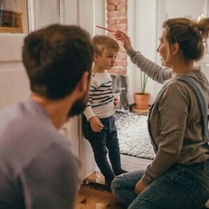 criança baixinha entenda a curva de crescimento infantil A mãe está ajoelhada checando o crescimento no filho, que está encostado na parede. Ela segura um lápis sobre a cabeça da criança para marcar a altura. O pai olha para a cena, um pouco mais distante.