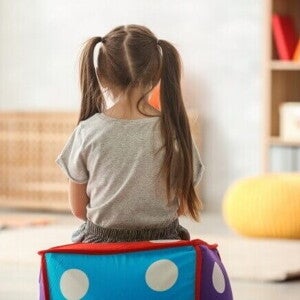 Criança sentada em cubo colorido em sala de terapia infantil, vista de costas.
