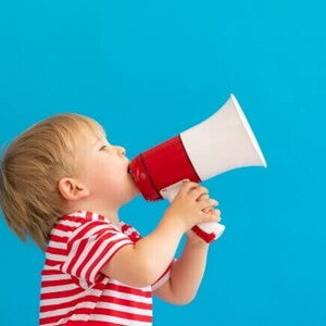 Um menino loiro usando camiseta listrada segura um megafone vermelho e branco contra um fundo azul sólido.