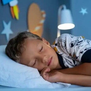 Criança dormindo Um menino, vestido de pijama estampado, está deitado na cama, de olhos fechados. O abajur atrás dele está aceso, e nas paredes do quarto há decorações de planetas.