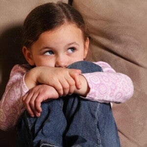 Uma criança medrosa sentada em um sofá marrom, abraçando os próprios joelhos com força e escondendo parte do rosto. Ela veste uma blusa rosa de manga longa e calça jeans, olhando para o lado com uma expressão de insegurança e apreensão.