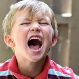 Close-up de uma criança gritando com a boca bem aberta e olhos fechados, expressando uma intensa explosão emocional ou crise de raiva.