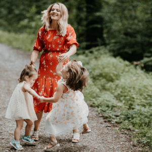 mulher, sorrindo e dançando com duas meninas pequenas em um campo, ambas vestindo vestidos brancos.