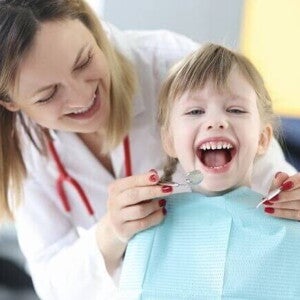 Criança sentada com a boca aberta, ao lado de uma dentista que está em pé, sorrindo e segurando aparelhos odontológicos. Criança sentada com a boca aberta, ao lado de uma dentista que está em pé, sorrindo e segurando aparelhos odontológicos.