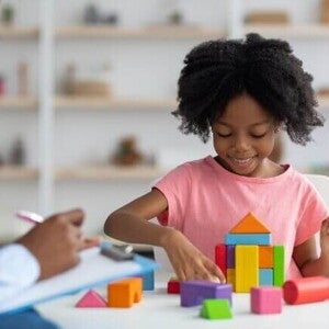 Uma menina sorrindo e organizando blocos coloridos, ao lado de uma mulher sentada com uma ficha na mão. Uma menina sorrindo e organizando blocos coloridos, ao lado de uma mulher sentada com uma ficha na mão.