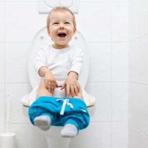 Uma criança sentada em um vaso sanitário com os shorts abaixados, rindo e olhando para cima, em um banheiro. Uma criança sentada em um vaso sanitário com os shorts abaixados, rindo e olhando para cima, em um banheiro.