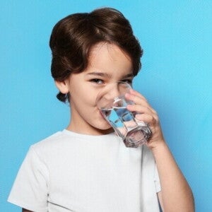Um menino está bebendo água de um copo transparente, olhando para a câmera, sob um fundo azul.