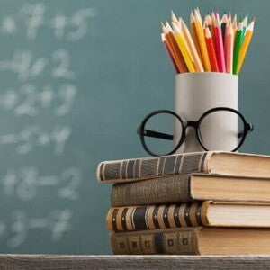 Uma pilha de livros sob uma mesa sustenta um par de óculos e uma caneca com lápis coloridos, com um quadro negro ao fundo Uma pilha de livros sob uma mesa sustenta um par de óculos e uma caneca com lápis coloridos, com um quadro negro ao fundo
