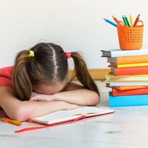 Menina deitada sobre um caderno aberto com a cabeça entre os braços, demonstrando exaustão e dificuldade de aprendizagem diante de uma pilha de livros e materiais escolares.