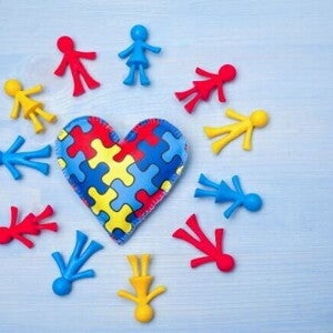 Coração feito de tecido com estampa de quebra-cabeça azul, vermelho e amarelo, simbolizando o autismo, cercado por pequenos bonecos coloridos de plástico.