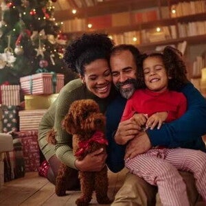 Homem e mulher abraçados com a filha e um cachorrinho no chão, na sala de casa em frente a uma árvore de natal Homem e mulher abraçados com a filha e um cachorrinho no chão, na sala de casa em frente a uma árvore de natal