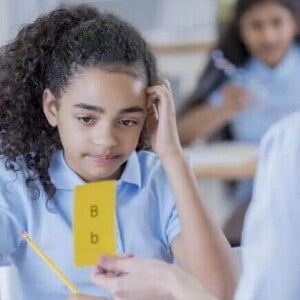Dislexia infantil Imagem de uma criança com dislexia olhando para uma ficha amarela com letras B maiúscula e minúscula. Em uma mão ela segura um lápis e a outra está sobre a cabeça em uma postura de dúvida.