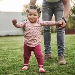 Imagem de um bebê cuja criação segue a abordagem de Emmi Pikler e que está brincando em um quintal supervisionado pela mãe que está lhe dando a mão. Imagem de um bebê cuja criação segue a abordagem de Emmi Pikler e que está brincando em um quintal supervisionado pela mãe que está lhe dando a mão.