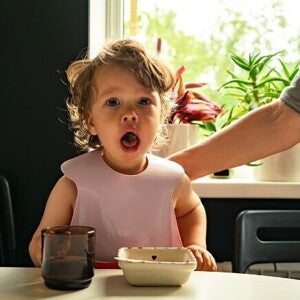 Um bebê com babador sentado à mesa, com a boca aberta, olhando diretamente para a câmera, com a mão de um adulto ao fundo.