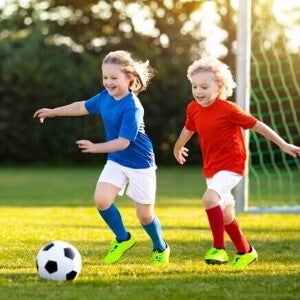 Duas crianças jogando futebol em um campo gramado sob a luz do sol, representando a prática de esportes para crianças.