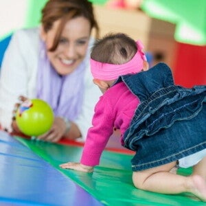 Um bebê engatinhando em direção a uma mulher sorridente, que observa o bebê com uma bola verde na mão.