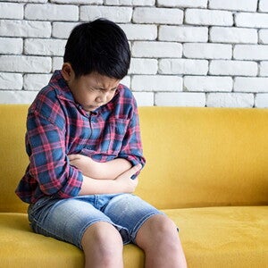 Estomatite infantil: o que é? Causas, sintomas e tratamento menino sentado no sofá com feição de dor abraçando a barriga