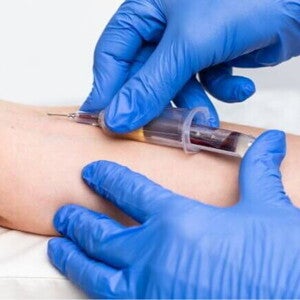 Profissional usando luvas azuis realizando coleta de sangue no braço de uma pessoa com uma seringa.