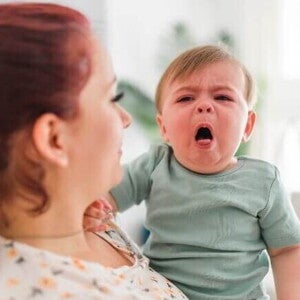 Mãe segurando um bebê que chora, com a boca aberta, indicando dor ou desconforto, possivelmente devido à faringite em bebês ou outra inflamação na garganta.