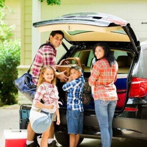 Família posando e sorrindo para a câmera, enquanto organiza malas dentro de um carro preto para sair de viagem