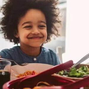 criança sentada a mesa no horário de almoço, sorrindo enquanto segura o talher na mão. Em sua frente estão os pratos com alimentos, incluindo uma salada verde, e há a presença de um adulto do outro lado da mesa.