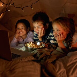 Três crianças, sendo um menino e duas meninas, deitados assistindo no notebook enquanto comem pipoca. Ao fundo, luminárias em uma cabana. Três crianças, sendo um menino e duas meninas, deitados assistindo no notebook enquanto comem pipoca. Ao fundo, luminárias em uma cabana.