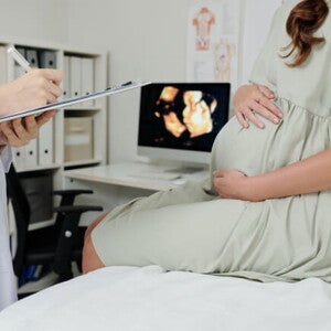 Gestante em consultório médico, sentada sob uma maca de atendimento, juntamente com um médico Gestante em consultório médico, sentada sob uma maca de atendimento, juntamente com um médico