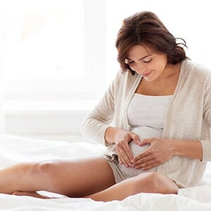 Imagem de uma gestante sentada na cama fazendo o formato de um coração sobre a sua barriga