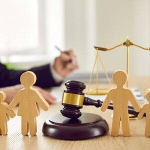 Objetos de direito sobre uma mesa de madeira, incluindo um martelo de juiz e bonecos de madeira representando uma família. A balança da justiça aparece ao fundo, e a imagem, com um juiz escrevendo ao fundo, reflete um processo judicial para a decisão de como funciona a guarda unilateral. Objetos de direito sobre uma mesa de madeira, incluindo um martelo de juiz e bonecos de madeira representando uma família. A balança da justiça aparece ao fundo, e a imagem, com um juiz escrevendo ao fundo, reflete um processo judicial para a decisão de como funciona a guarda unilateral.
