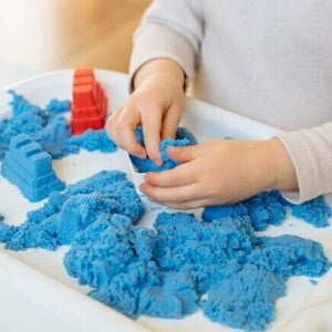 Close das mãos de uma criança interagindo com areia cinética azul em uma bandeja branca.