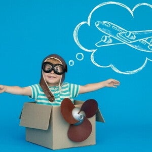 Um menino usando óculos de aviador e um capacete de couro decola de dentro de uma caixa de papelão, sorrindo. Acima de sua cabeça, uma bolha de pensamento branca sobre um fundo azul vibrante mostra um desenho de um avião. Um menino usando óculos de aviador e um capacete de couro decola de dentro de uma caixa de papelão, sorrindo. Acima de sua cabeça, uma bolha de pensamento branca sobre um fundo azul vibrante mostra um desenho de um avião.