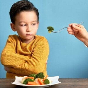 Uma mão estendendo um garfo com brócolis para um menino, que olha de forma irritada para o alimento. Uma mão estendendo um garfo com brócolis para um menino, que olha de forma irritada para o alimento.