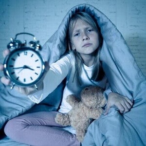 Uma menina loira, com uma expressão de cansaço ou preocupação, sentada na cama coberta por um edredom, segurando um despertador à sua frente, com um ursinho de pelúcia ao lado.