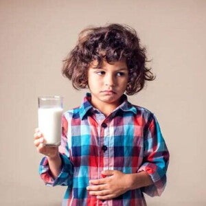 Intolerância à lactose em crianças: como identificar? Imagem de uma criança desconfortável, com um copo de leite em uma mão e outra sobre a barriga em uma postura que remete aos efeitos da intolerância à lactose.