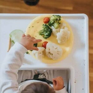 Introdução alimentar Uma fotografia tirada de cima mostra um bebê em fase de introdução alimentar e seu prato amarelo com vegetais coloridos a sua frente