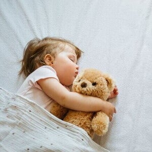 Uma bebê deitada em uma cama, dormindo, abraçada com um ursinho de pelúcia. Uma bebê deitada em uma cama, dormindo, abraçada com um ursinho de pelúcia.