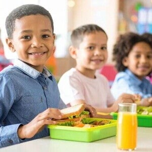 Crianças felizes e sorridentes, comendo sanduíches e suco durante o recreio, utilizando uma lancheira verde cheia de alimentos saudáveis como vegetais e frutas.