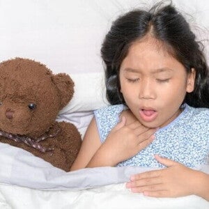Uma menina deitada na cama com expressão de desconforto, segurando a garganta, ao lado de um ursinho de pelúcia. Uma menina deitada na cama com expressão de desconforto, segurando a garganta, ao lado de um ursinho de pelúcia.