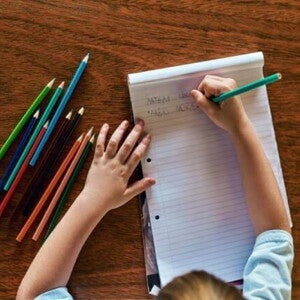 Um menino está escrevendo em um bloco de folhas alguns nomes, como “Fabio”. Na mesa estão espalhados lápis de cor de diversas cores. A foto está ligada ao tema Troca de letras na infância.
