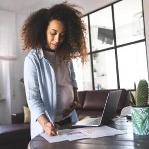 Uma mulher grávida escreve em alguns papéis na mesa da sala de casa, enquanto está de licença-maternidade. 